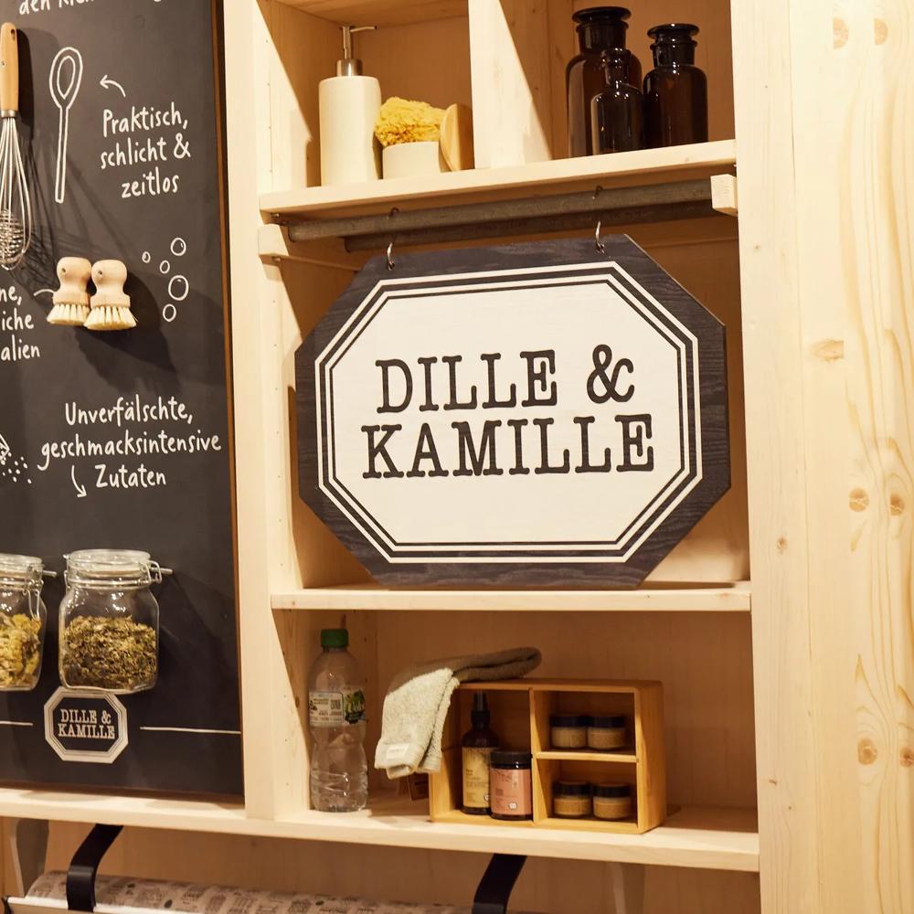 Leinwandtaschen und ein Schild mit "Dille & Kamille" an einer Holzwand, daneben Gläser mit Kräutern.