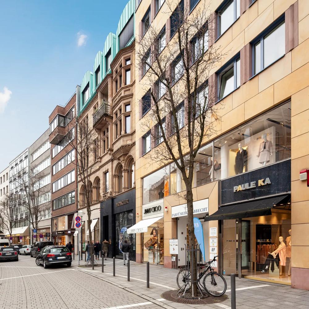 Geschäftsstraße mit Autos, Fahrrädern und Fußgängern, gesäumt von modernen und historischen Gebäuden.