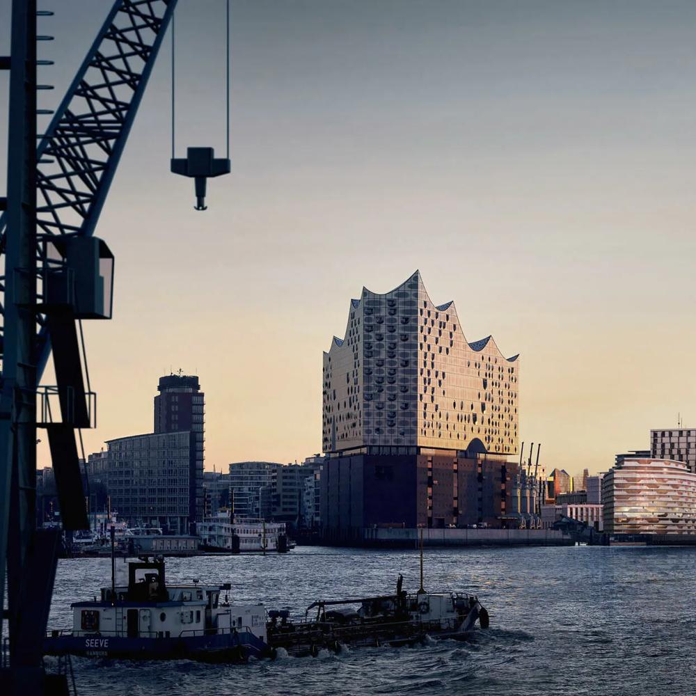 Ein modernes Gebäude mit wellenförmigem Dach am Wasser bei Sonnenuntergang, Kran und Schiff im Vordergrund.
