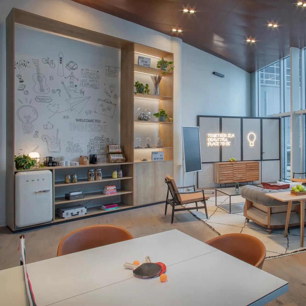 Moderner Raum mit Sofa, Tisch, roten Hockern, Pflanzen und großen Fenstern. Wandregale und Tischtennisschläger im Vordergrund.