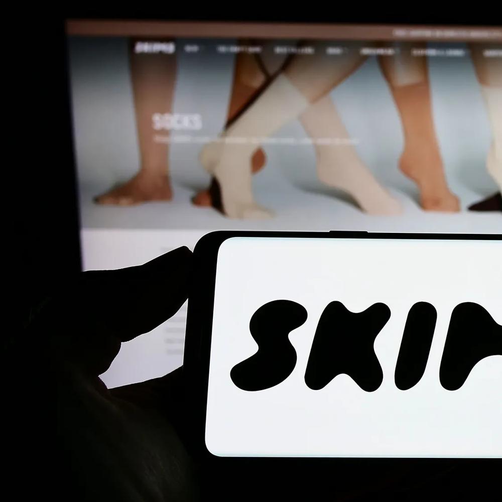 Smartphone mit Skims-Logo vor einer Website mit Produktdarstellung auf einem Bildschirm.