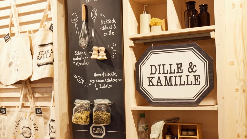 Leinwandtaschen und ein Schild mit "Dille & Kamille" an einer Holzwand, daneben Gläser mit Kräutern.