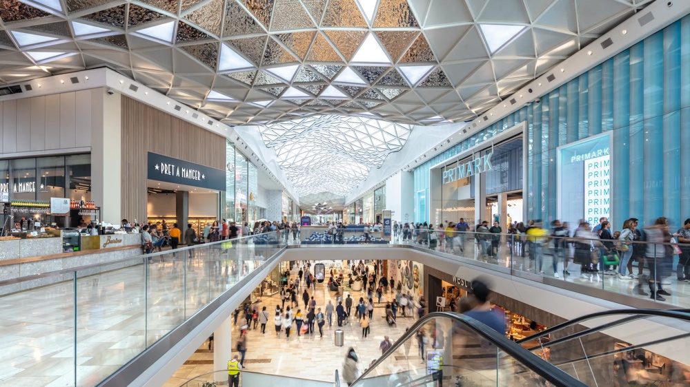 Menschenmengen in einem modernen Einkaufszentrum mit Rolltreppen und Geschäften auf mehreren Ebenen.