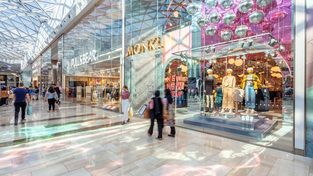 Einkaufszentrum mit gläsernem Dach, Geschäften und Schaufensterpuppen in bunter Beleuchtung, Menschen flanieren.