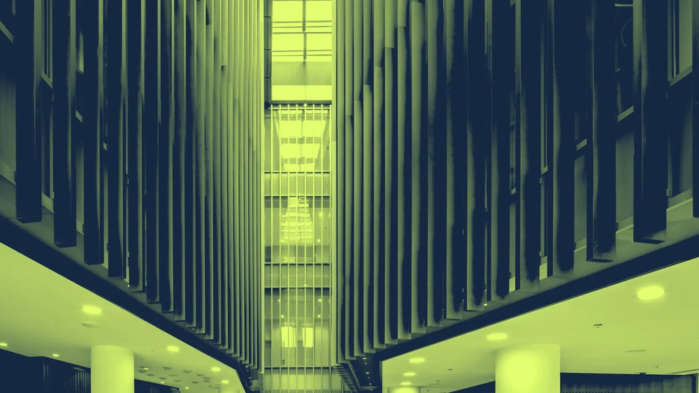 Moderner Innenraum mit hohen Lamellenwänden und Deckenlichtern, zentraler Glasaufzug im Hintergrund.