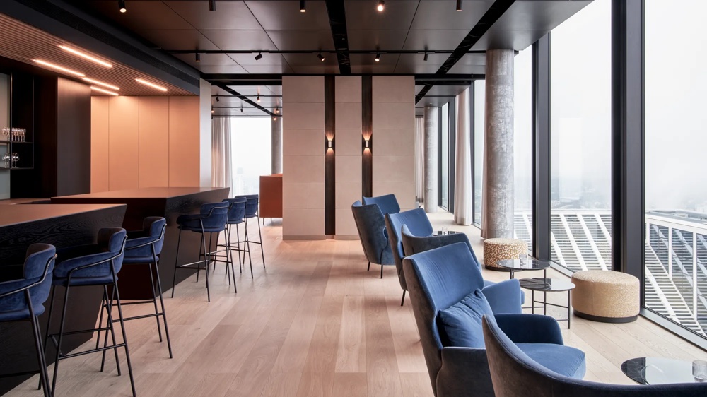 Moderne Lounge mit blauen Stühlen, Barhockern und großen Fenstern mit Stadtblick. Helle Holzböden und schwarze Decke.