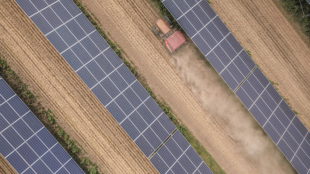 Ein Traktor pflügt ein Feld zwischen Reihen von Solarpanels aus der Vogelperspektive.