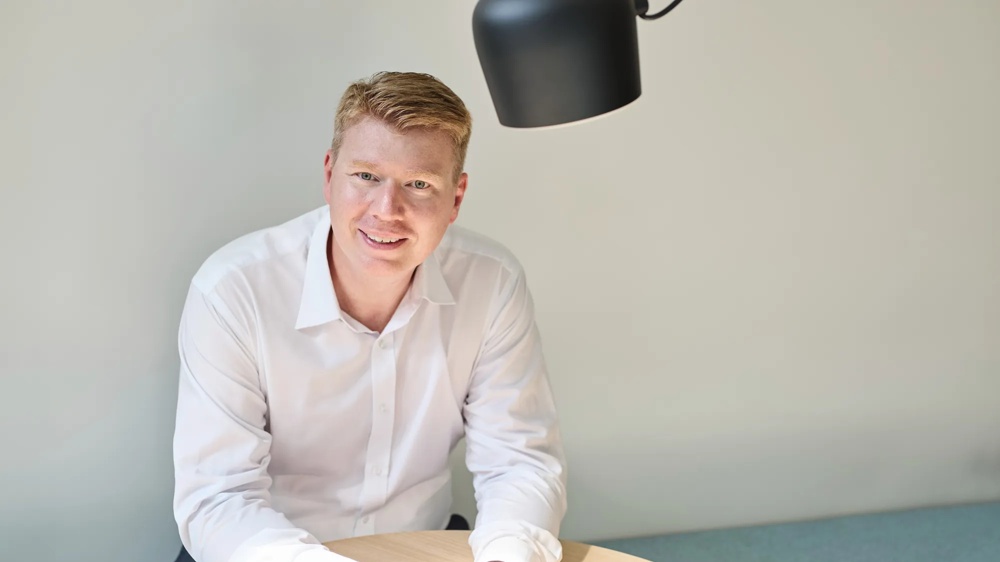 Eine Person in weißem Hemd sitzt lächelnd an einem runden Holztisch unter einer schwarzen Lampe.