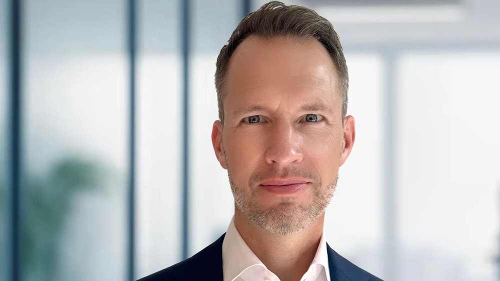 A man in a suit is standing in front of a blurry, modern background.