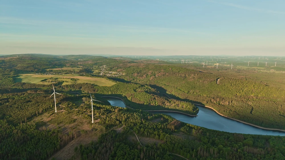 Windräder auf einem grünen Hügel unter bewölktem Himmel mit Sonnenstrahlen.