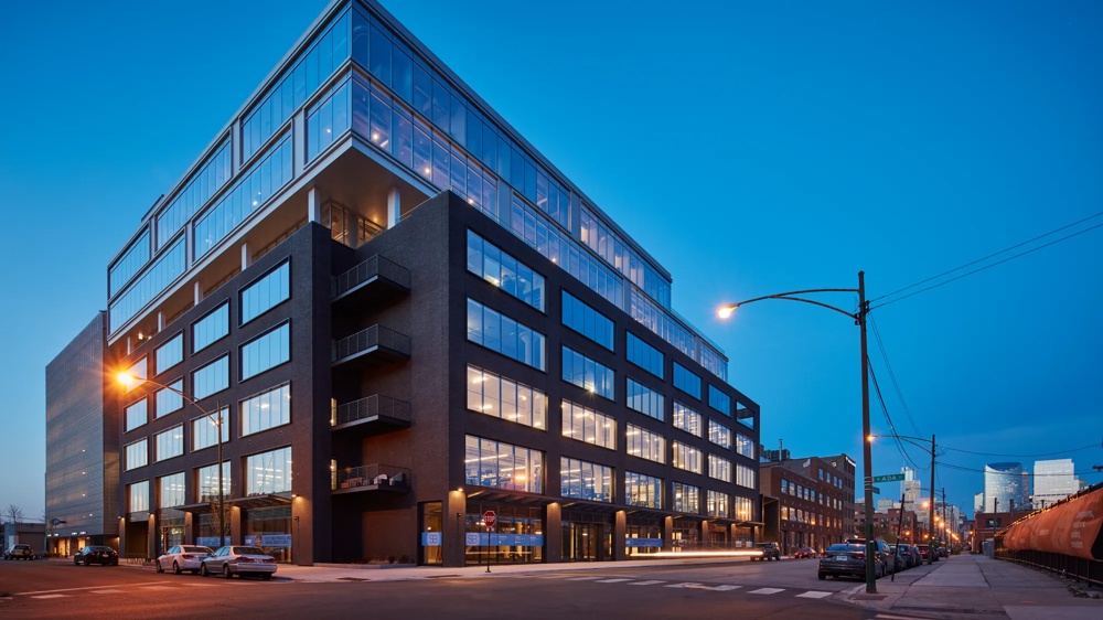 Modernes Bürogebäude bei Dämmerung, beleuchtete Fenster, Straßenlaternen, blauer Himmel, Autos am Straßenrand.