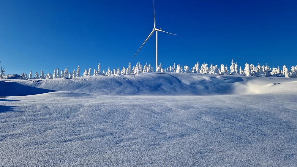 A windmill stands in a snowy landscape against a clear, blue sky, surrounded by snow-covered trees.