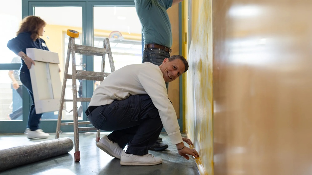 Zwei Männer renovieren eine gelbe Wand, eine Frau trägt ein weißes Brett, Leiter im Hintergrund.