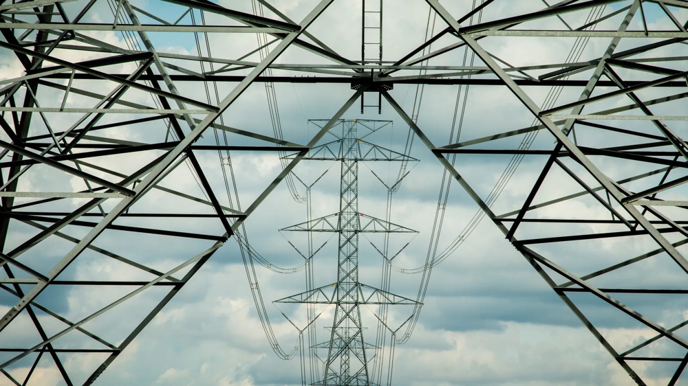 Blick durch das Stahlgerüst eines Hochspannungsmastes auf einen weiteren Mast im Hintergrund vor bewölktem Himmel.