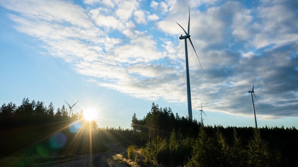 Windräder stehen auf einem Feld unter blauem Himmel mit Wolken, während die Sonne am Horizont aufgeht.
