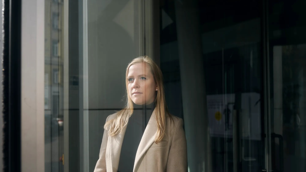 Eine Frau im beigen Mantel steht hinter einer Glasscheibe, blickt nachdenklich hinaus.