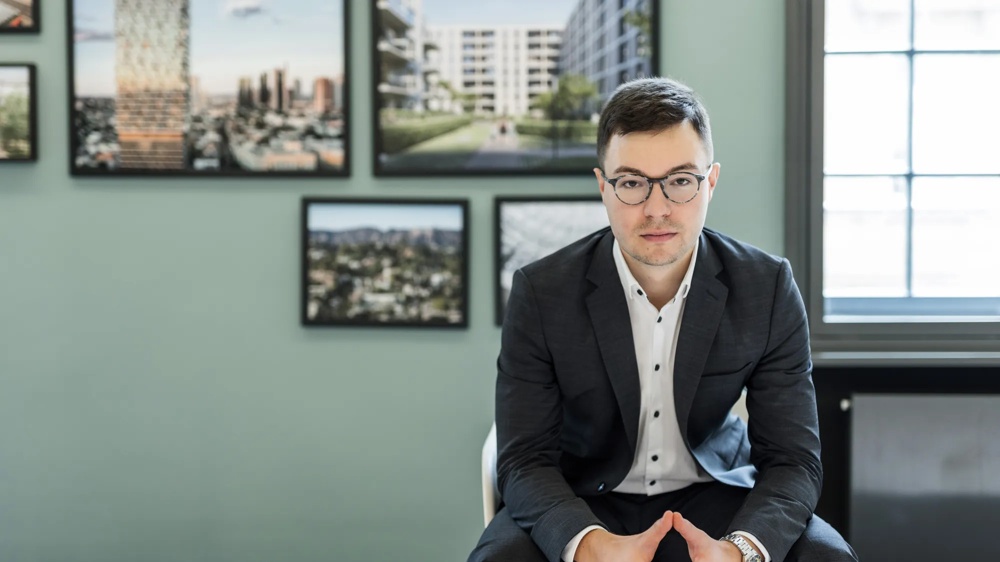 Ein Mann im Anzug sitzt vor einer grauen Wand mit gerahmten Bildern.