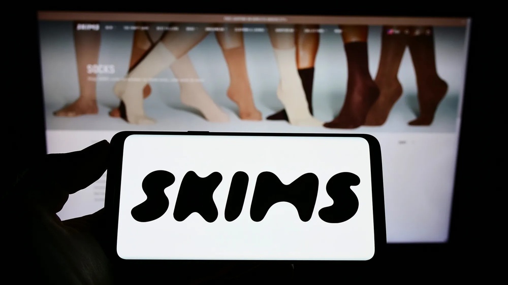 Smartphone mit Skims-Logo vor einer Website mit Produktdarstellung auf einem Bildschirm.