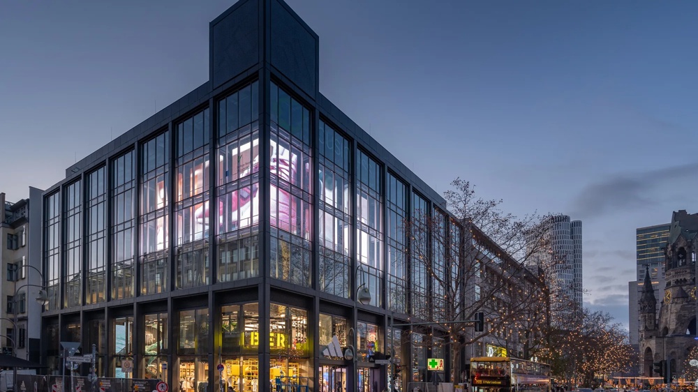 Modernes Glasgebäude bei Dämmerung, leuchtende Werbeanzeigen, Menschen und Bus auf belebter Straße.