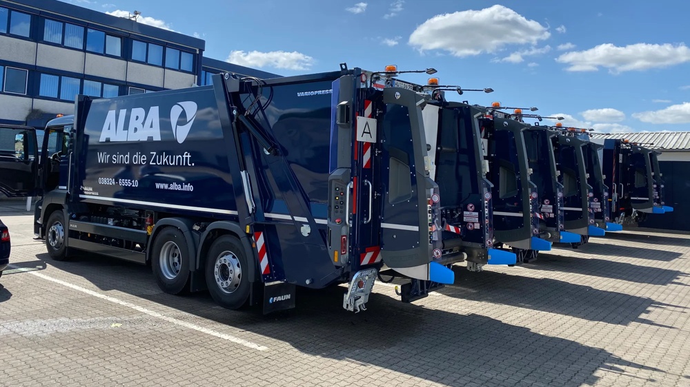 Eine Reihe von blauen Müllwagen auf einem gepflasterten Parkplatz bei sonnigem Wetter.