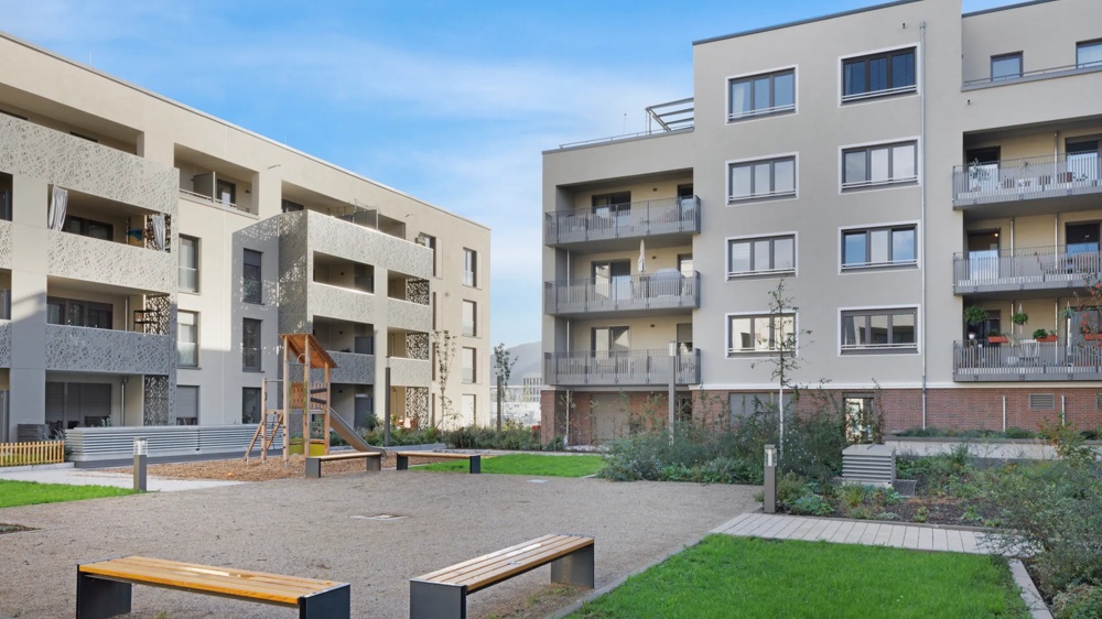 Moderne Wohngebäude mit Balkonen, Spielplatz und Bänken in einem grünen Innenhof unter blauem Himmel.