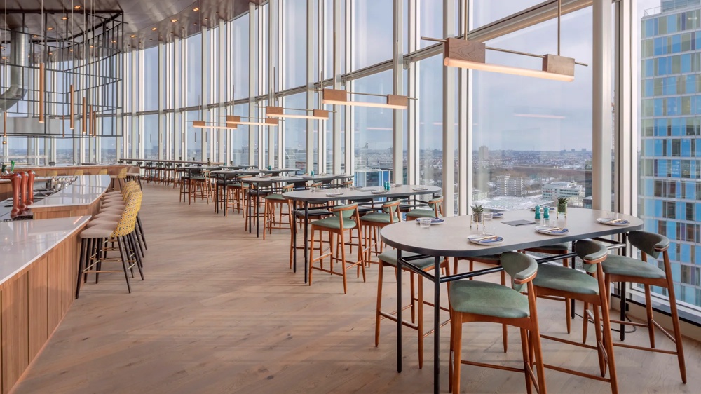 Ein modernes Restaurant mit hohen Fenstern, Holztischen und grünen Stühlen, bietet einen weiten Stadtblick.