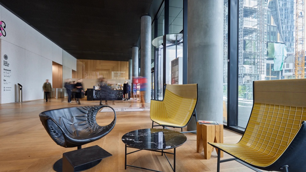 Moderne Lobby mit gelben Sesseln, rundem Marmortisch und hohen Glasfenstern, Menschen im Hintergrund, Holzfußboden.