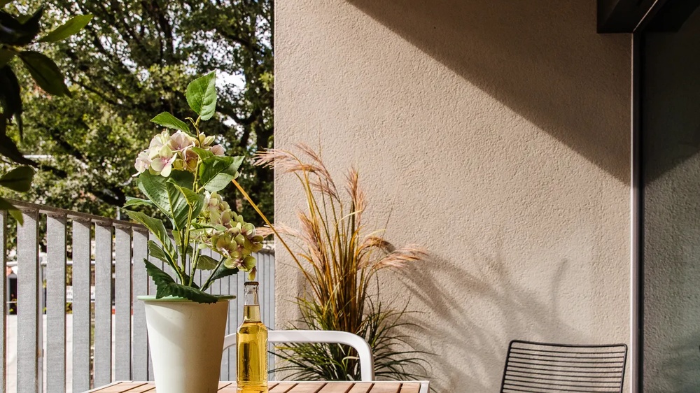 Ein Balkon mit Tisch, Pflanze, Getränk, Stuhl und Decke, von Sonnenlicht beschienen, mit Bäumen im Hintergrund.