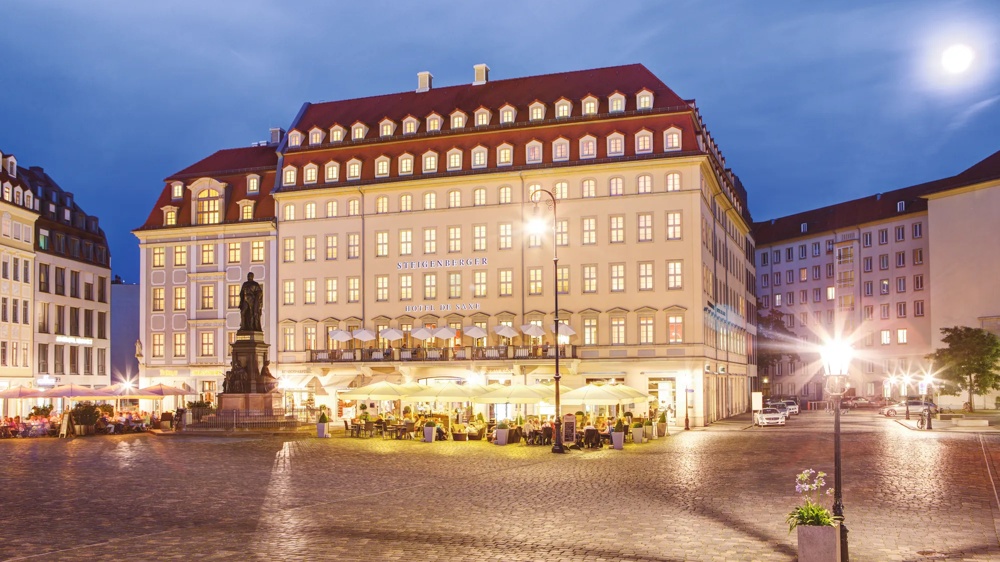 An illuminated hotel building on the square at dusk, with people in a café and a statue in the foreground.