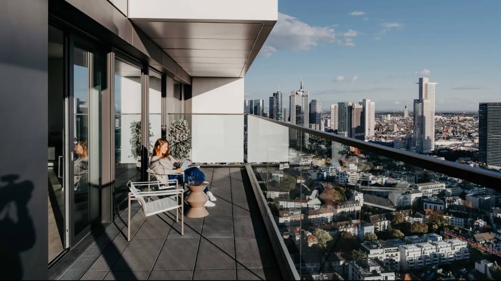 Woman sitting on a balcony with a view of the city, reading in the sunshine, skyscrapers in the background.