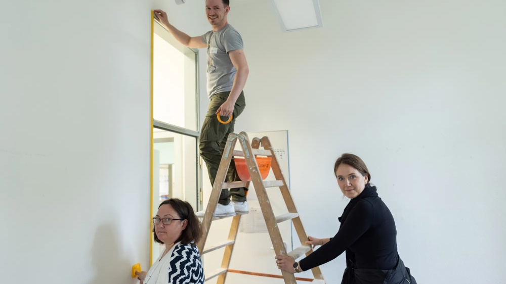 Drei Personen arbeiten an einer Leiter, umrahmen eine Tür mit gelbem Klebeband in einem weißen Raum.