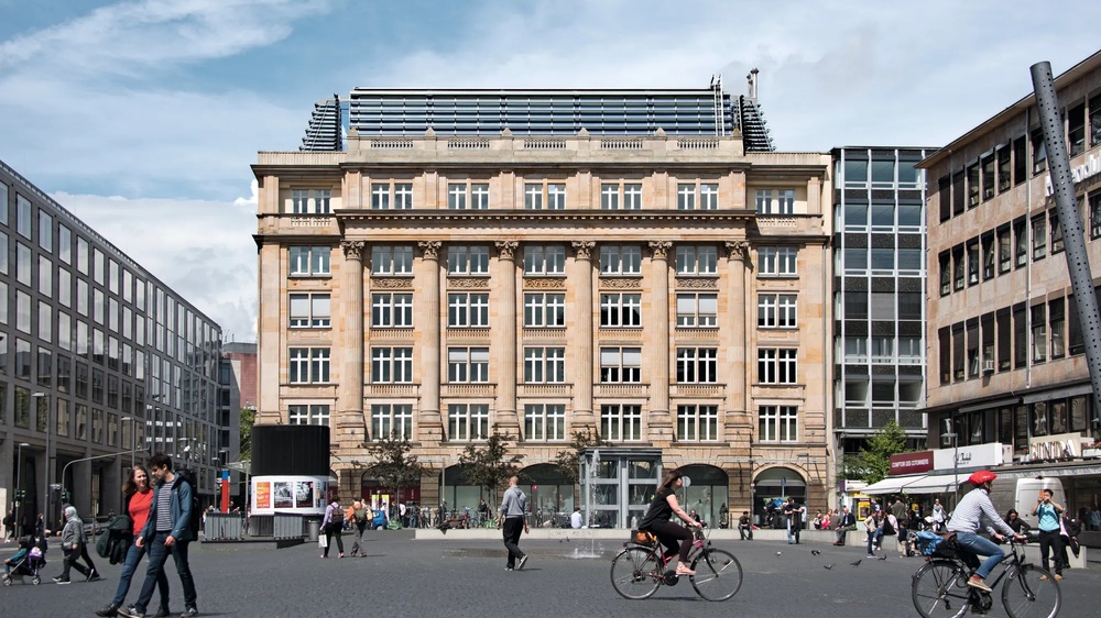 Ein historisches Gebäude mit vielen Fenstern, davor Menschen zu Fuß und auf Fahrrädern auf einem belebten Platz.
