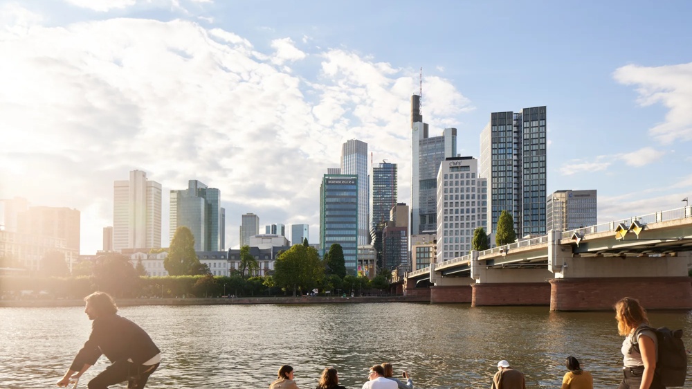 Radfahrer und Spaziergänger am Flussufer, Skyline und Brücke im Hintergrund.