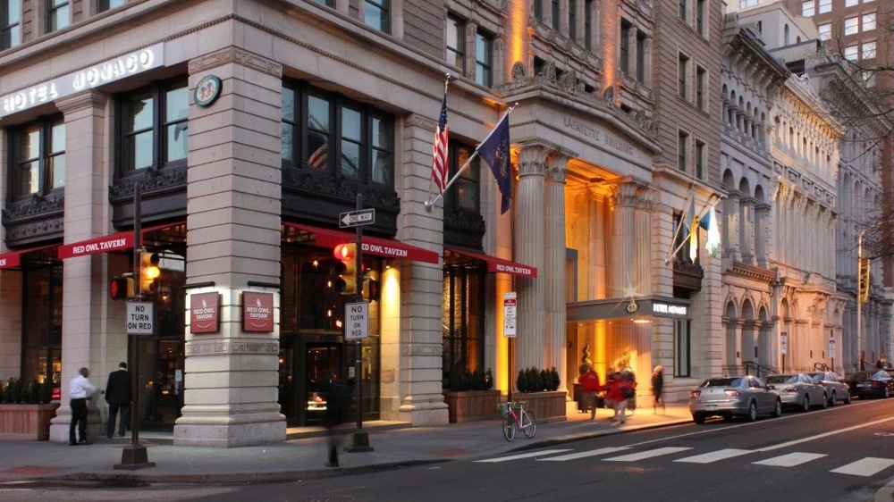 Eckgebäude mit Hotel und Restaurant, beleuchtet, mit Flaggen und Fußgängern auf der Straße bei Abenddämmerung.