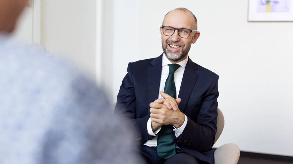 Ein lächelnder Mann in Anzug und Krawatte sitzt auf einem Stuhl in einem Büro.
