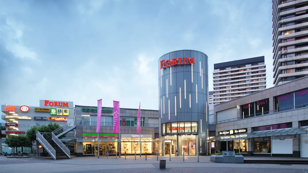 Einkaufszentrum mit grauem Turm, rotem "Forum" Schriftzug, umgeben von Geschäften und hohen Gebäuden.