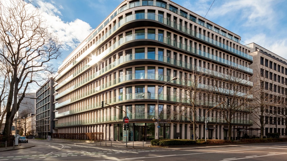 Modern office building with a glass facade at a street corner, blue sky and bare trees in the foreground.