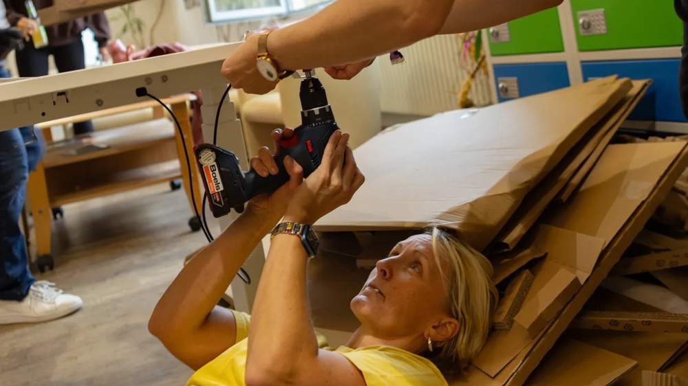Person montiert Tisch mit Bohrmaschine, liegt auf Pappe. Andere helfen, bunte Schränke im Hintergrund.
