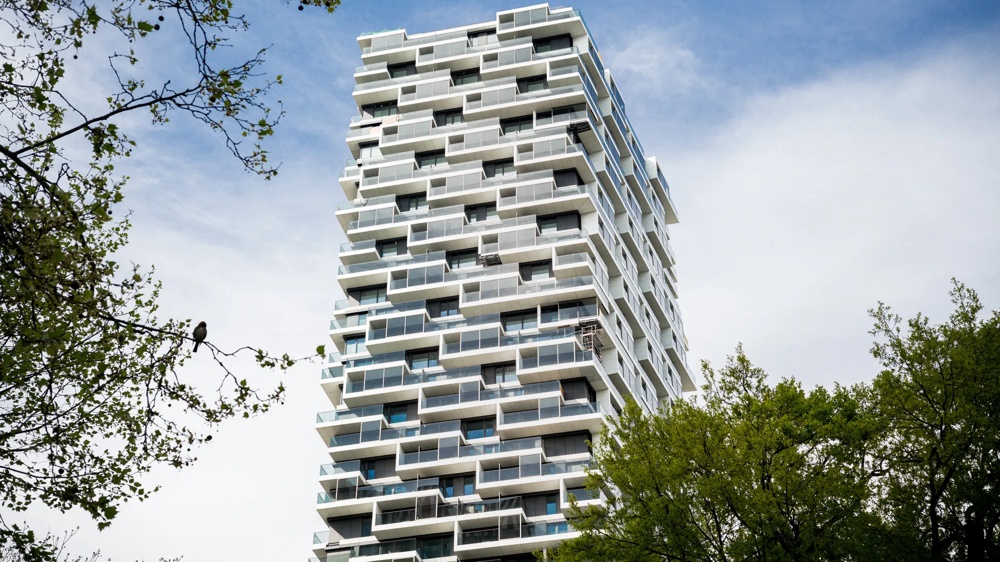 A modern high-rise with offset balconies, surrounded by green trees and a blue sky.