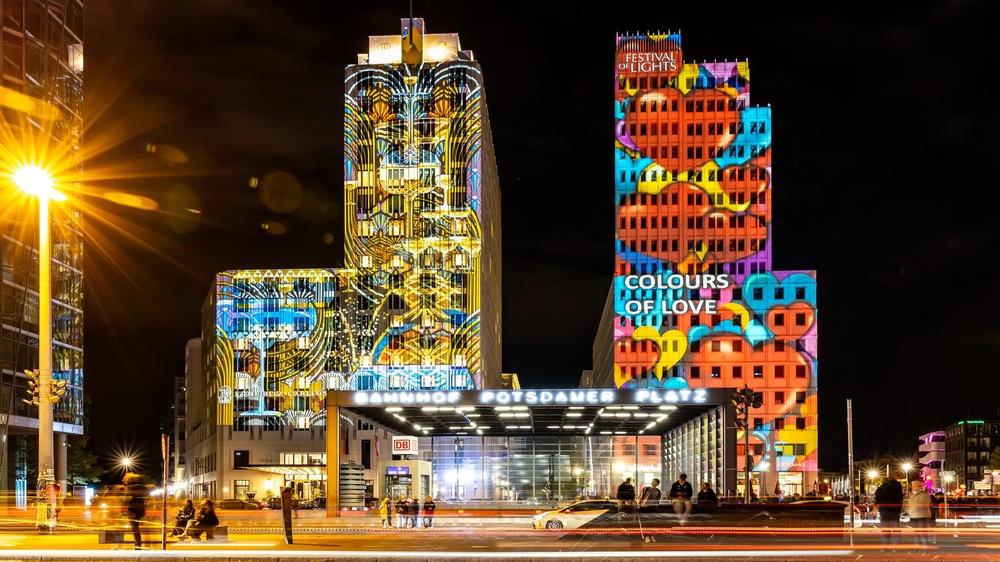 Mehrgeschossiges Gebäudeensemble bei Nacht mit großflächigen farbigen Lichtprojektionen auf den Fassaden.