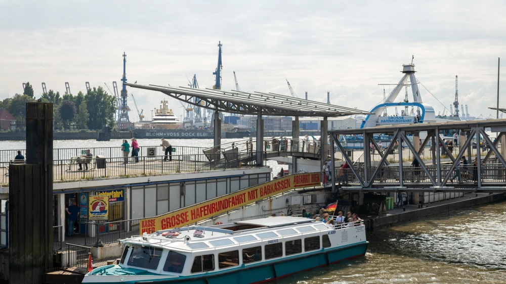 Ein Boot liegt an einem Hafenanleger, im Hintergrund Kräne und ein großes Schiff.
