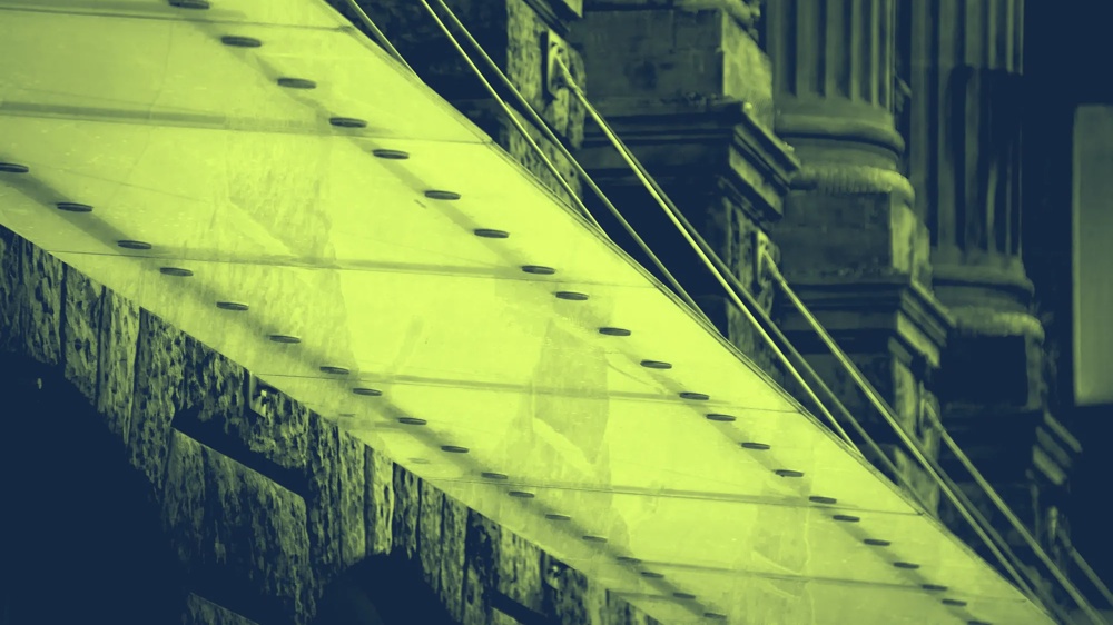 Glasüberdachung mit Metallseilen vor alten Steinsäulen in gelb-grünem Farbton.