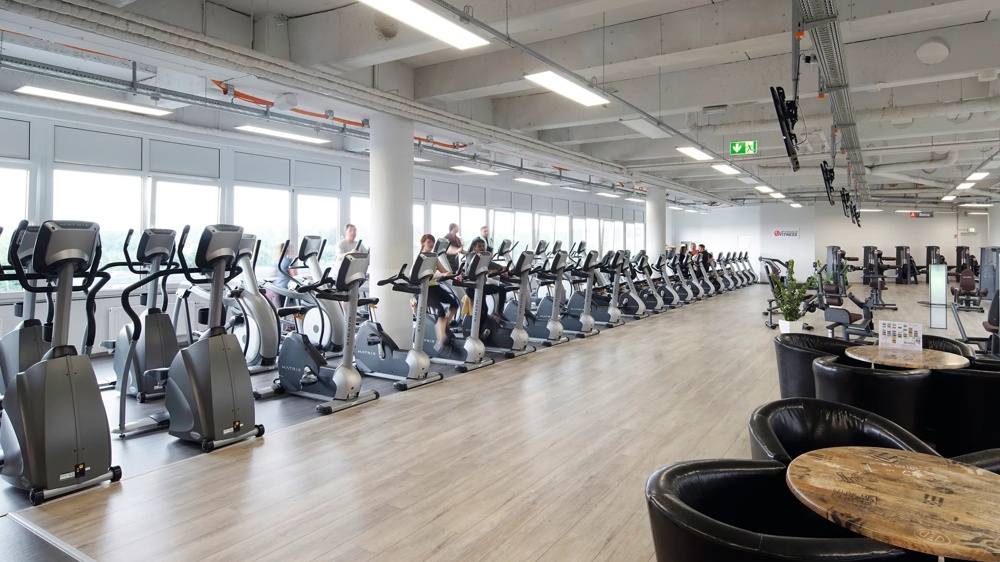 Reihe von Fitnessgeräten in einem hellen Fitnessstudio, Menschen trainieren auf Crosstrainern.