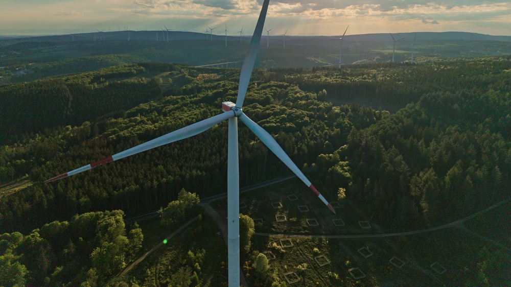 Sonnenuntergang über einem Wald mit Windrädern am Horizont und Wolken am Himmel.