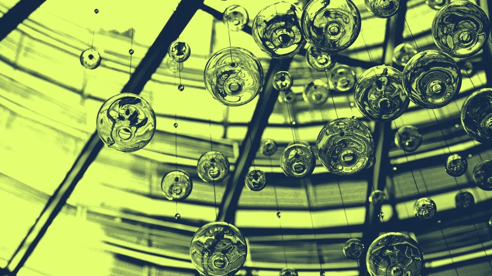 Glasblasen hängen an Fäden vor einem geometrischen, grün getönten Dachfenster.