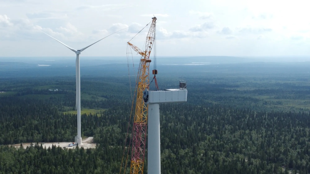Zwei Windräder stehen im Wald und eines davon wird mithilfe eines Krans fertig zusammengesetzt