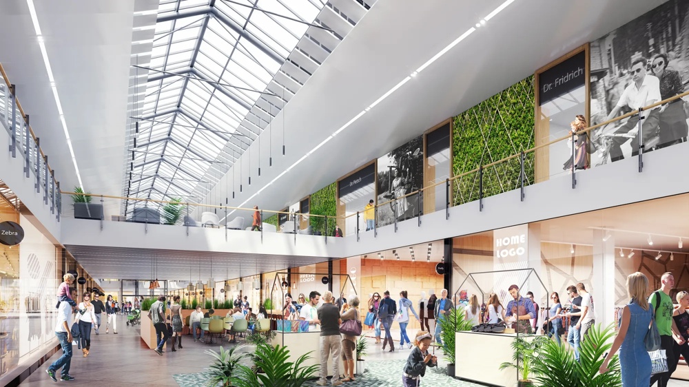 Helle Einkaufspassage mit Glasdach, Menschen flanieren, Pflanzen und Geschäfte auf beiden Seiten.