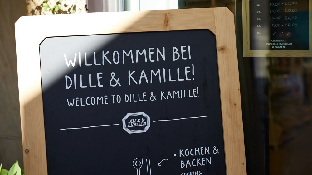 Willkommensschild von Dille & Kamille neben einem Fenster mit Öffnungszeiten und Pflanzen im Hintergrund.