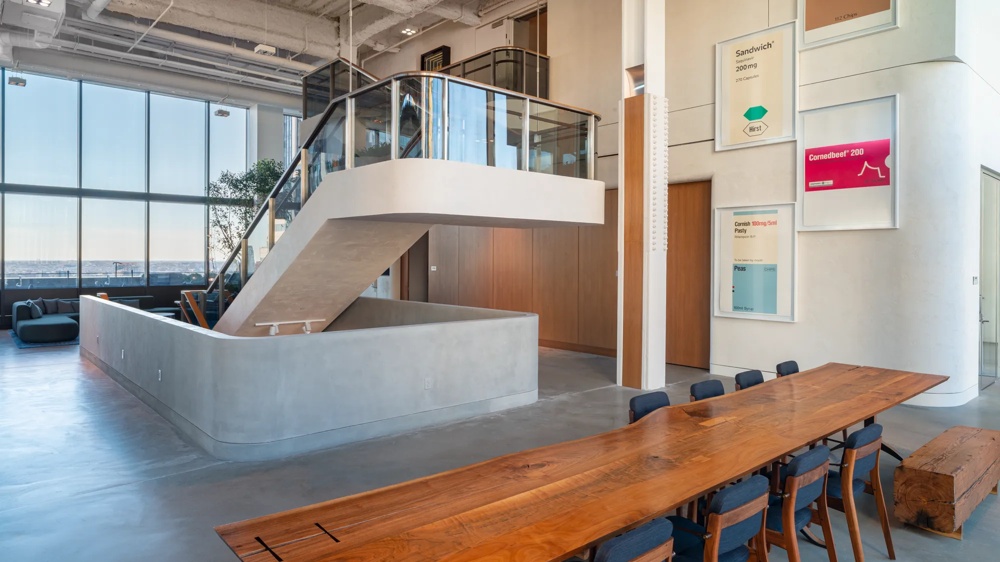 Moderner Büroraum mit Holztisch, blauen Stühlen, Treppe, großen Fenstern und Wandbildern.