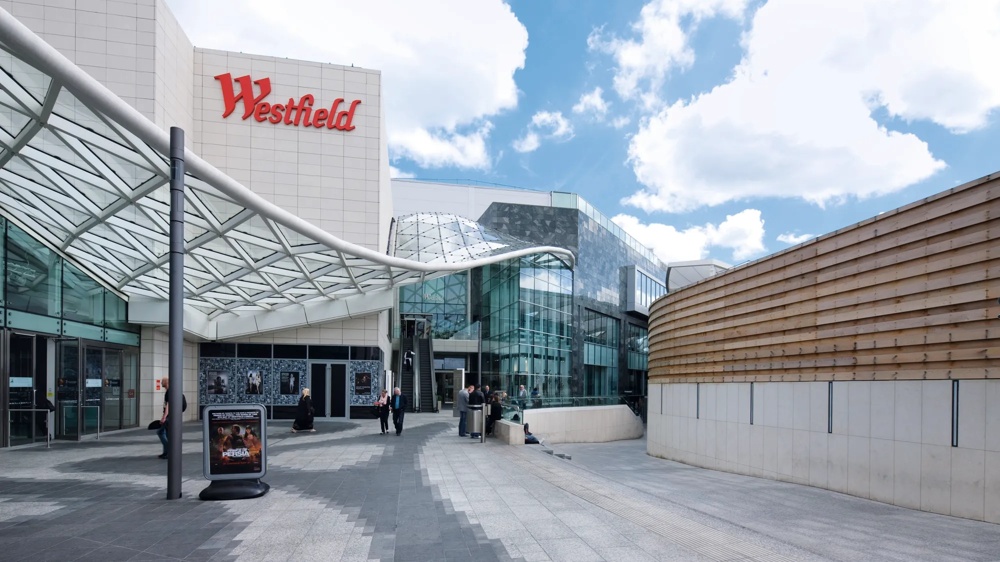Eingang eines modernen Einkaufszentrums mit Glasdach und Westfield-Logo unter blauem Himmel.